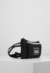 Svart PUMA midjeväska med en texturerad yta, framficka med dragkedja och justerbar rem med tryckt logotyp. Kompakt design för bekvämlighet.