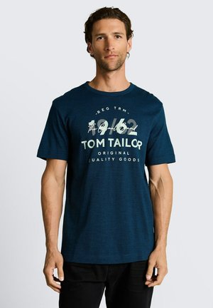 Mann trägt ein marineblaues T-Shirt mit kurzen Ärmeln und dem Text „1962 TOM TAILOR Original Quality Goods“, steht vor einem schlichten, hellen Hintergrund.