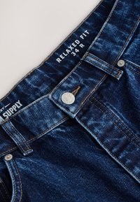 Tmavomodré riflové nohavice so zapínaním na gombíky, s pútkami na opasok a bielym textom vo vnútri pásu s nápisom "RELAXED FIT 34 R."