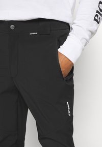 Un pantalon noir extensible avec une ceinture boutonnée, des poches latérales zippées et un détail de logo. Le matériau semble lisse avec une coupe ajustée.
