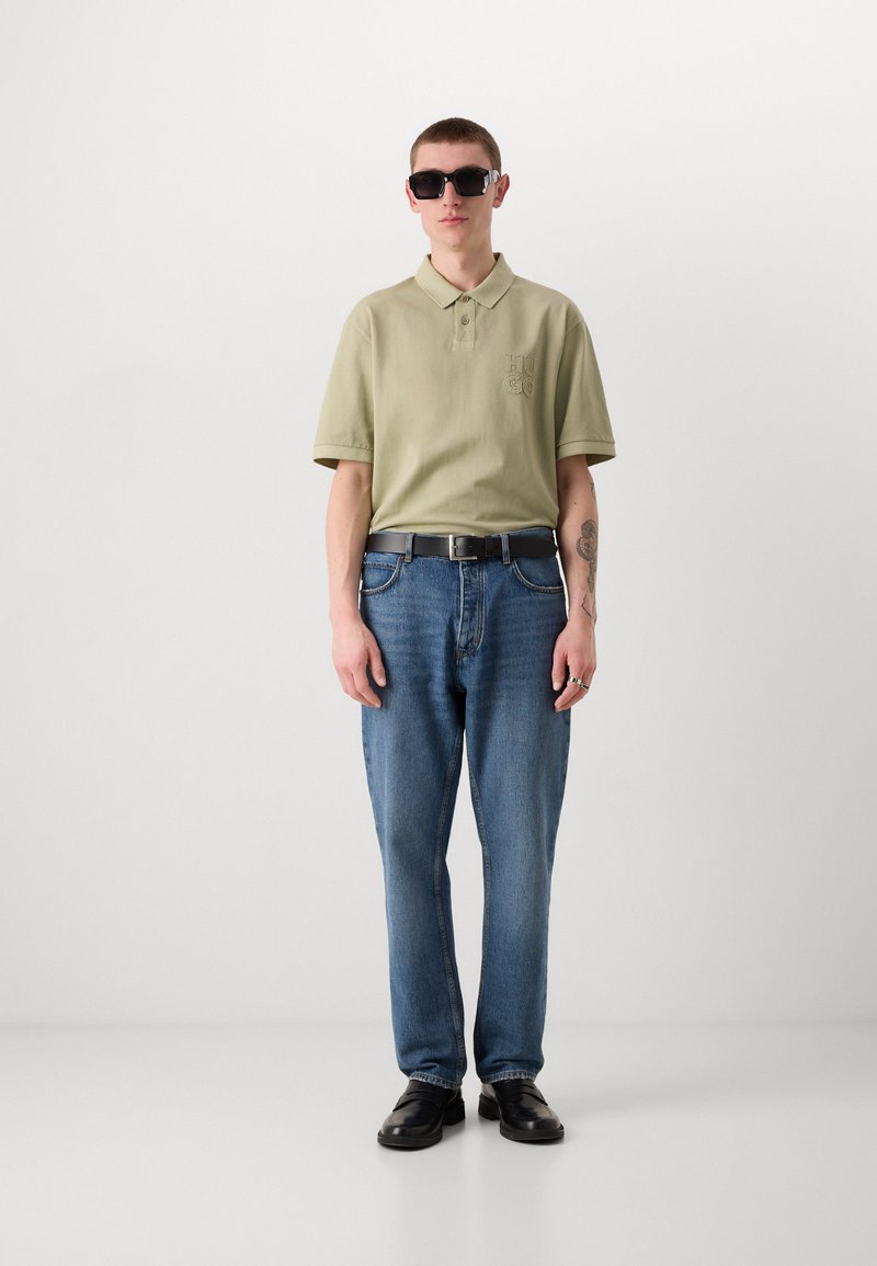 Polo vert clair avec col boutonné, logo brodé, jeans bleus, ceinture noire et lunettes de soleil noires avec monture rectangulaire.