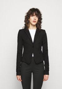 Blazer noir ajusté avec une fermeture à un bouton, des revers à cran, et deux poches avant, associé à un top blanc et un pantalon sombre.