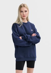 Navyblauwe sweatshirt met een ronde hals en geribbelde boorden. Heeft een klein geborduurd ontwerp op de borst. Draagt het met zwarte shorts.