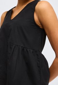 Κοντινό πλάνο ενός ατόμου που φοράει μαύρο αμάνικο τοπ με V-λαιμόκοψη και λεπτομέρεια πιέτας κάτω από το στήθος.