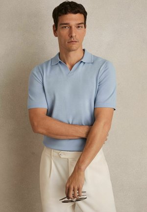 Hellblaues Kurzarm-Polohemd mit V-Ausschnitt, aus weichem Stoff; kombiniert mit cremefarbenen Hosen und hält eine Sonnenbrille.
