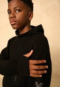 Haut à capuche noir tricoté avec une texture côtelée et des poignets avec trou pour le pouce. Il comporte une poche avant et une étiquette sur la manche. Le mannequin pose les bras croisés.