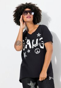 Camiseta negra de manga corta con letras blancas "GAGE" y acentos de estrellas plateadas, que presenta un diseño de cuello en v y un ajuste relajado.