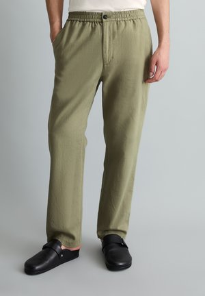 Man draagt olijfgroene broek met elastische taille en zwarte instapschoenen, staand met handen gedeeltelijk in de zakken.