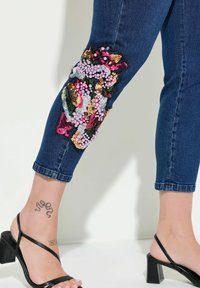 Tummansiniset farkku-ankle-farkut, joissa on värikkäitä paljettikoristeita polvialueella. Ominaisuuksiin kuuluu vartalonmyötäinen muotoilu ja taitettu helma. Mustat hihnalliset sandaalit.