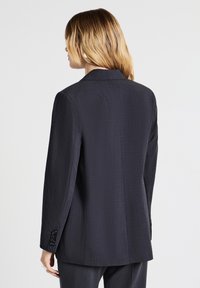 Donna con capelli biondi alla spalla, che indossa un blazer grigio scuro su misura e pantaloni abbinati, ripresa da dietro.