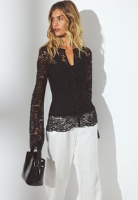 Black lace blouse with long sleeves, button-up front, scalloped hem, worn over a black top, paired with white linen pants and a black handbag.