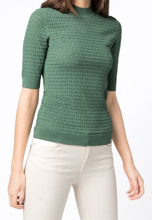 Femme portant un haut en tricot vert ajusté à manches courtes avec un motif texturé et un pantalon couleur crème, sur fond blanc.