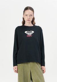 Long-sleeve black t-shirt with a white heart logo and pink "ROXY" text. Smooth fabric, relaxed fit, crew neckline, worn with green corduroy pants.