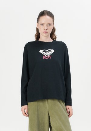 Long-sleeve black t-shirt with a white heart logo and pink "ROXY" text. Smooth fabric, relaxed fit, crew neckline, worn with green corduroy pants.
