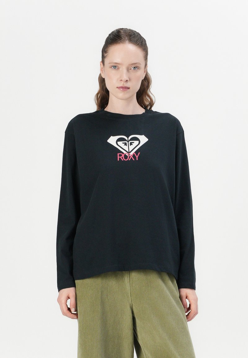 Long-sleeve black t-shirt with a white heart logo and pink "ROXY" text. Smooth fabric, relaxed fit, crew neckline, worn with green corduroy pants.