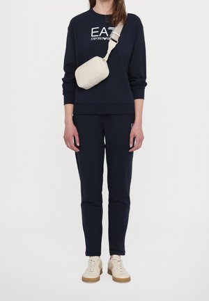 Persona che indossa una felpa e pantaloni blu navy Emporio Armani, una borsa a tracolla beige e scarpe bianche, in piedi davanti a uno sfondo neutro.