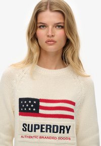 Gebreide crèmekleurige trui met een ontwerp van de Amerikaanse vlag in rood en blauw, met het woord "SUPERDRY" in marineblauwe tekst eronder. Geweven patroon door de hele trui.