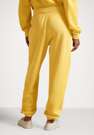 Rumene športne hlače iz mehke tkanine z elastičnim pasom, rebrastimi manšetami in sproščenim krojem. Noser obut v svetle čevlje.