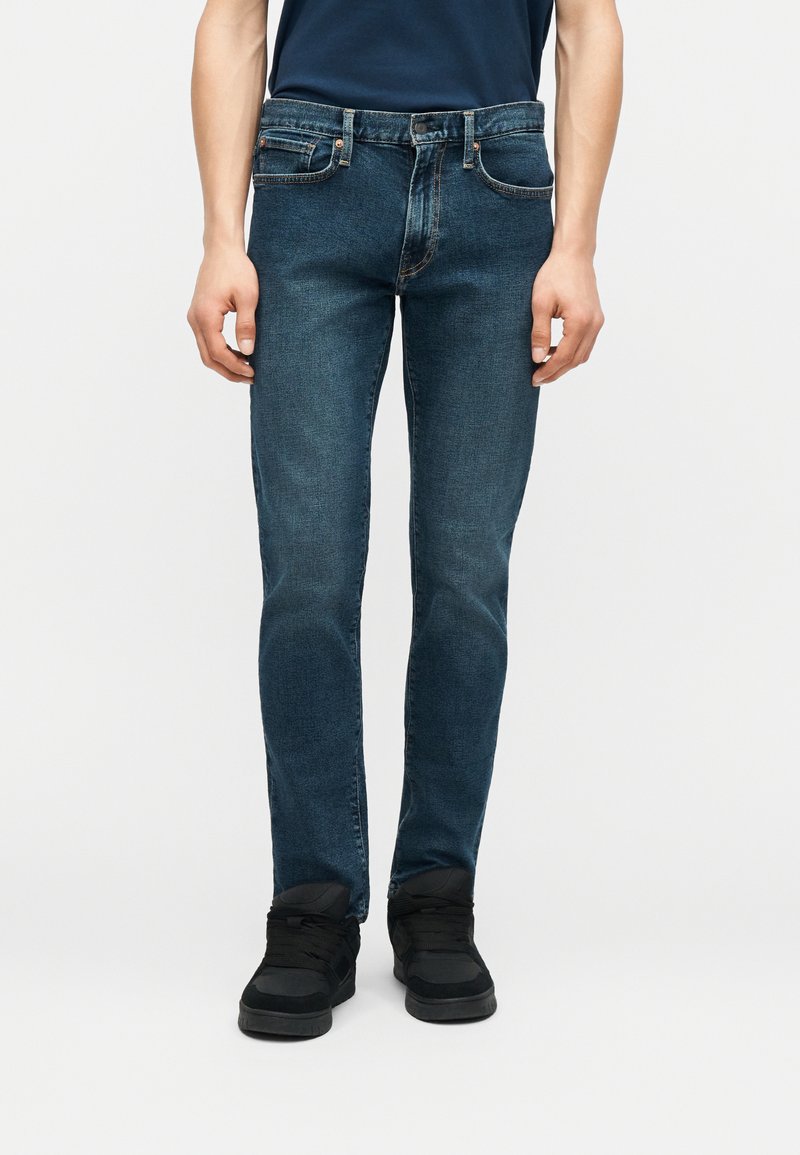 Homme portant un jean slim bleu foncé et des baskets noires, debout devant un fond clair uni, les mains détendues le long du corps.