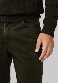 Pantalon en velours côtelé vert foncé avec une coupe slim, présentant des côtes verticales et une poche avant. Un pull en tricot à côtes est partiellement visible au-dessus.