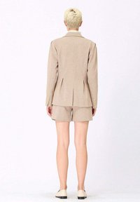 Blazer à carreaux beige avec shorts assortis, texture lisse, coupe ajustée et plis arrière discrets. Des chaussures de couleur claire complètent la tenue.