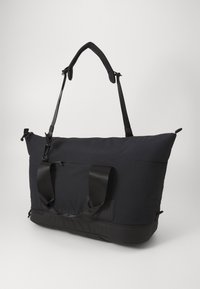 Black fabric duffel bag with adjustable shoulder straps, top handles, and a textured surface. Features a zippered outer pocket and logo detail.
