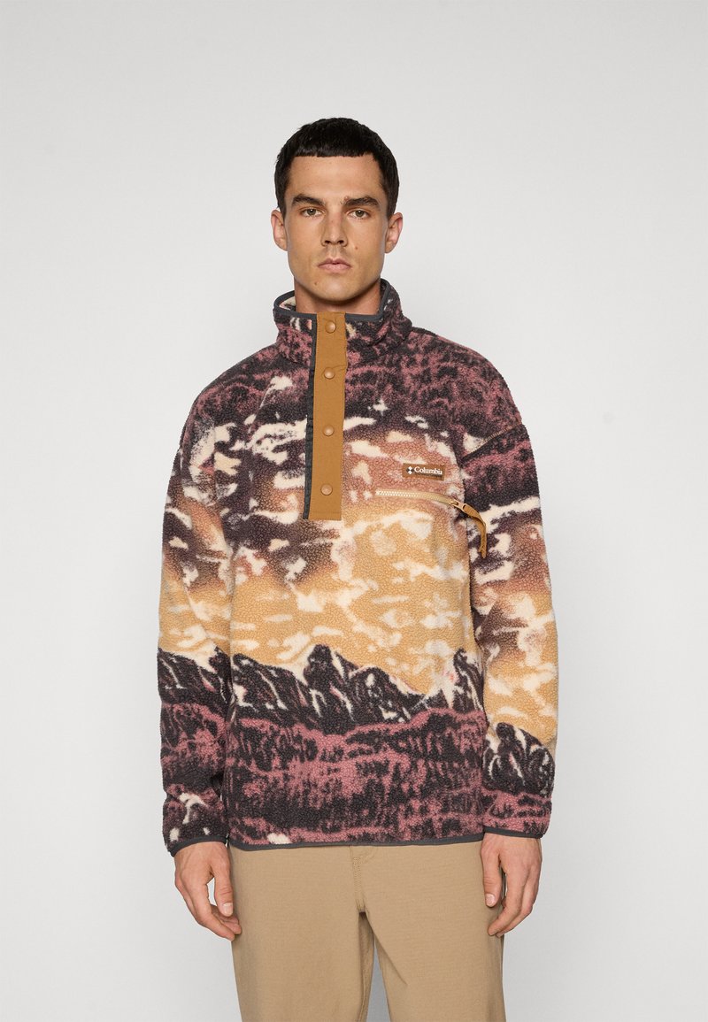 Man in een meerkleurige fleecepullover met bergpatronen, met knoopkraag, borstzak met rits en beige broek tegen een effen achtergrond.