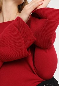 Femme portant un pull rouge côtelé avec de larges manches cloche superposées, croisant les bras près de son cou et de ses épaules.