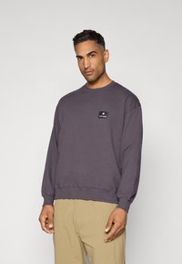 Felpa viola in cotone, con collo rotondo, maniche lunghe e una piccola etichetta logo nera sul petto. Texture liscia.