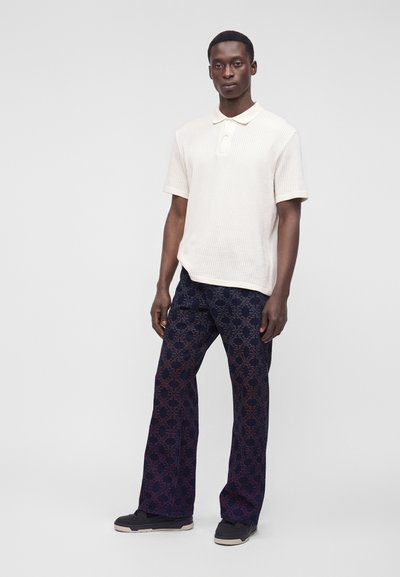 Homme portant un polo blanc à manches courtes, un pantalon large foncé à motifs et des baskets noires, debout devant un fond clair uni.