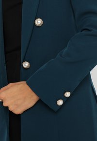 Marynarka w kolorze morskim o gładkiej fakturze, z dużymi perłowymi guzikami na mankietach i przodzie. Projekt zawiera klapy z wycięciami i dopasowany krój.