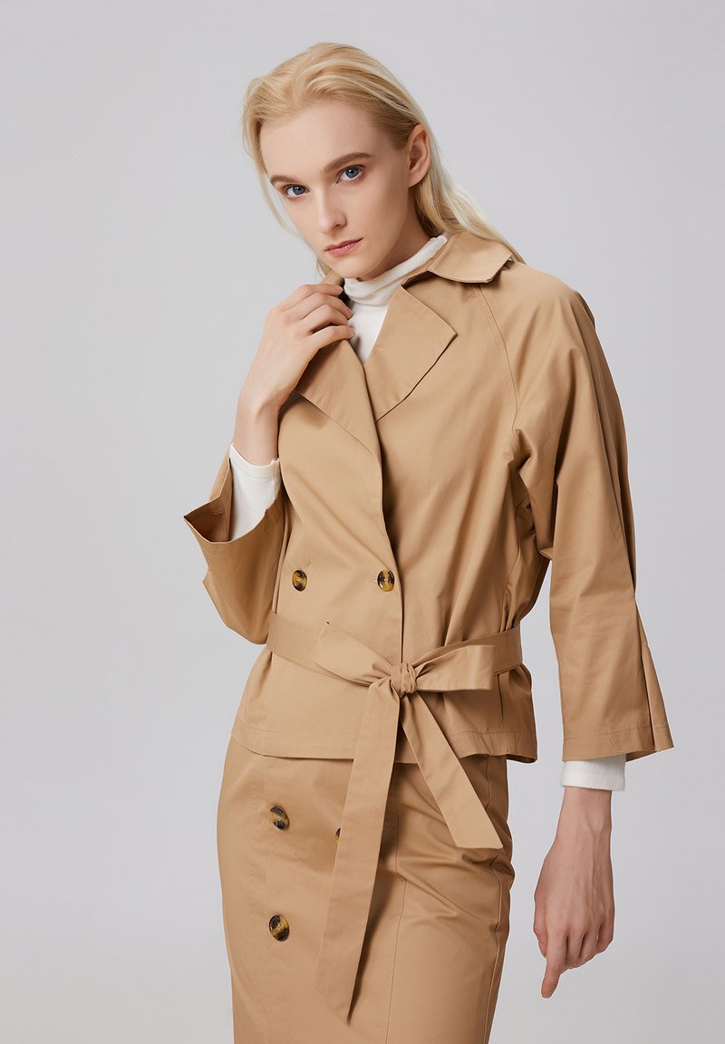 Veste beige croisée avec une ceinture à nouer, dotée d'un col, de manches longues et de boutons brillants. Texture de tissu lisse.