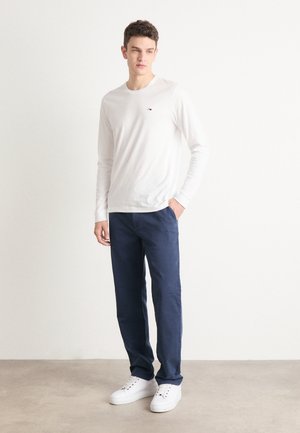 Camisa blanca de manga larga con un pequeño logo en el pecho, combinada con pantalones azul marino y zapatillas blancas. Tela suave, corte casual, diseño minimalista.