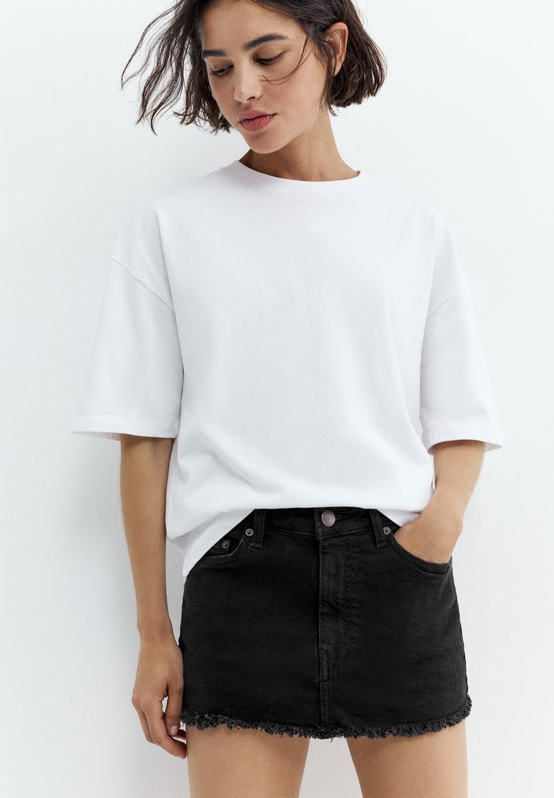Femme aux cheveux courts et foncés portant un t-shirt blanc ample et une mini-jupe en jean noire à ourlet effiloché, une main dans la poche, regardant vers le bas.