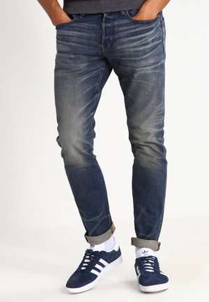 Mann trägt hochgekrempelte, blaue verwaschene Jeans und marineblaue Adidas-Sneaker mit weißen Socken, Hände in den Taschen vor einfachem Hintergrund.