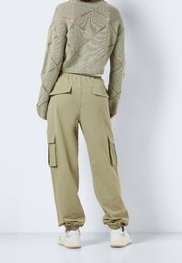 Grön kortärmad tröja med öppen stickning, tillsammans med ljusolivfärgade cargobyxor med fickor och elastiska muddar. Vita sneakers synliga.