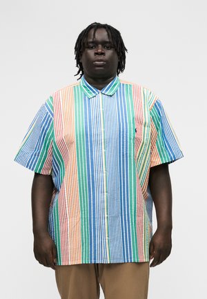 Νεαρός άνδρας με σκουρόχρωμο δέρμα και dreadlocks, φορώντας πολύχρωμο ριγέ πουκάμισο με κοντά μανίκια και μπεζ παντελόνι, σε απλό φόντο.