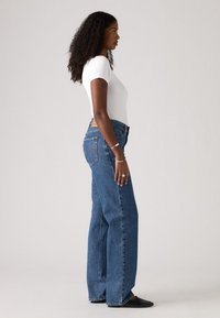 Hoge taille, wijde blauwe denim jeans met een vervaagde was, zijzakken en een klassiek vijfzakkenontwerp. G gedragen met een getailleerde witte top.