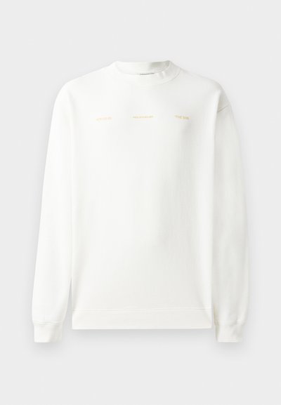 Valge dressipluus ümmarguse kaeluse, pikkade varrukate ja soonikuga mansetiga. Rinnal on kuldse trükiga tekst: "HZW (25-26) HOLZWEILER THE SUN."