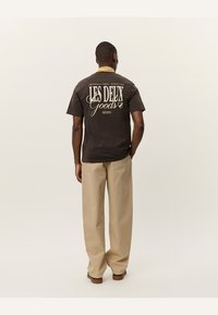 Camisa marrón de manga corta con un diseño de texto blanco, acentos en blanco y negro, y un corte relajado; combinada con pantalones de pierna ancha beige.