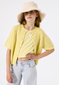 Cárdigan de punto amarillo claro con mangas cortas, combinado con una blusa a juego de canalé y jeans de pierna ancha azul claro. Sombrero tipo cubo beige y collar floral.