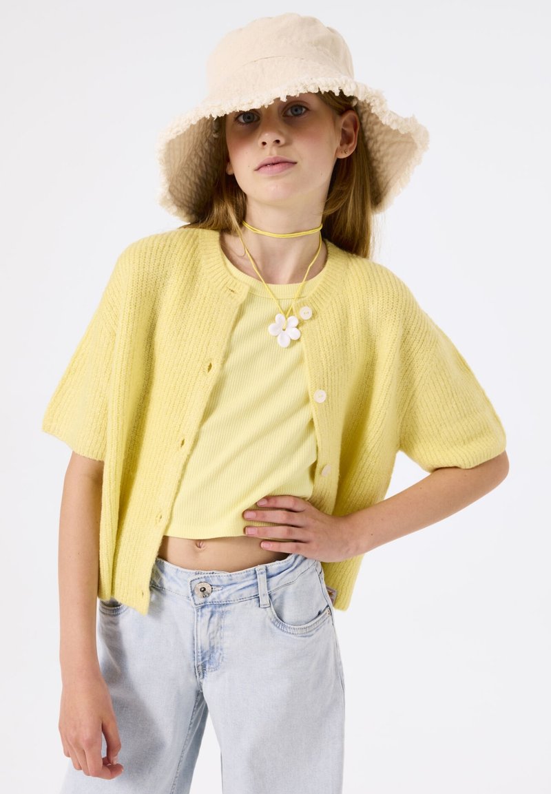 Cárdigan de punto amarillo claro con mangas cortas, combinado con una blusa a juego de canalé y jeans de pierna ancha azul claro. Sombrero tipo cubo beige y collar floral.