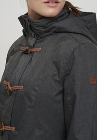 Grå jakke med et strukturert, fiskebensmønster, hette, treknapper og brune fletteakkenter. Logo-patch på ermet.