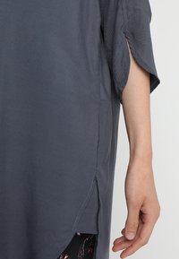 Szara, oversize bluzka z krótkim rękawem, z bocznymi rozcięciami i gładką teksturą. Materiał wydaje się lekki i ma swobodny styl.