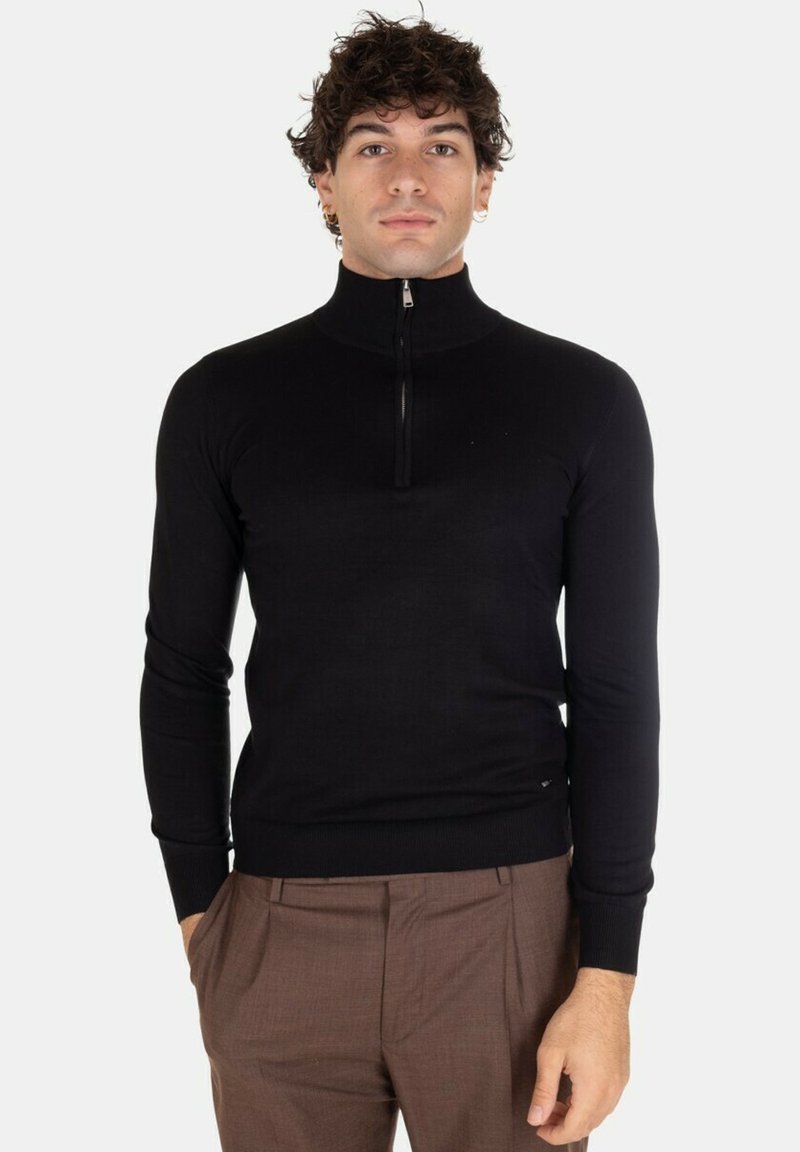 Maglione nero con zip al collo, maniche lunghe, polsini e orlo a coste, realizzato in tessuto liscio, design aderente, abbinato a pantaloni marroni.