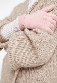 Strickhandschuhe in Hellrosa mit gerippten Bündchen. Die Hand lehnt sich an einen strukturierten beigen Pullover, der die weichen Materialien hervorhebt.