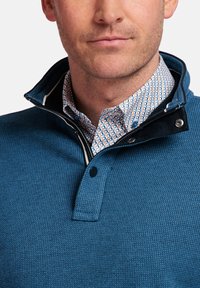 Blauw gestructureerde pullover met een hoge kraag, voorzien van een knoopsluiting en een patroonshirt eronder met cirkelvormige ontwerpen in oranje en blauw.