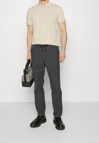 Homme portant un polo beige, un pantalon de jogging gris foncé avec cordon de serrage, des chaussures en cuir noires, tenant un sac de sport gris à motifs avec des poignées noires.