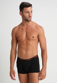 Musta bokserityyppinen alusvaate, jossa on oranssi logo-yläosa. Valmistettu sileästä kankaasta, istuvalla muotoilulla ja lyhyellä lahkeella.