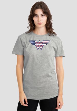 Woman with wavy brown hair wearing a gray t-shirt featuring a red, white, and blue "Wonder Woman" logo with American flag design.